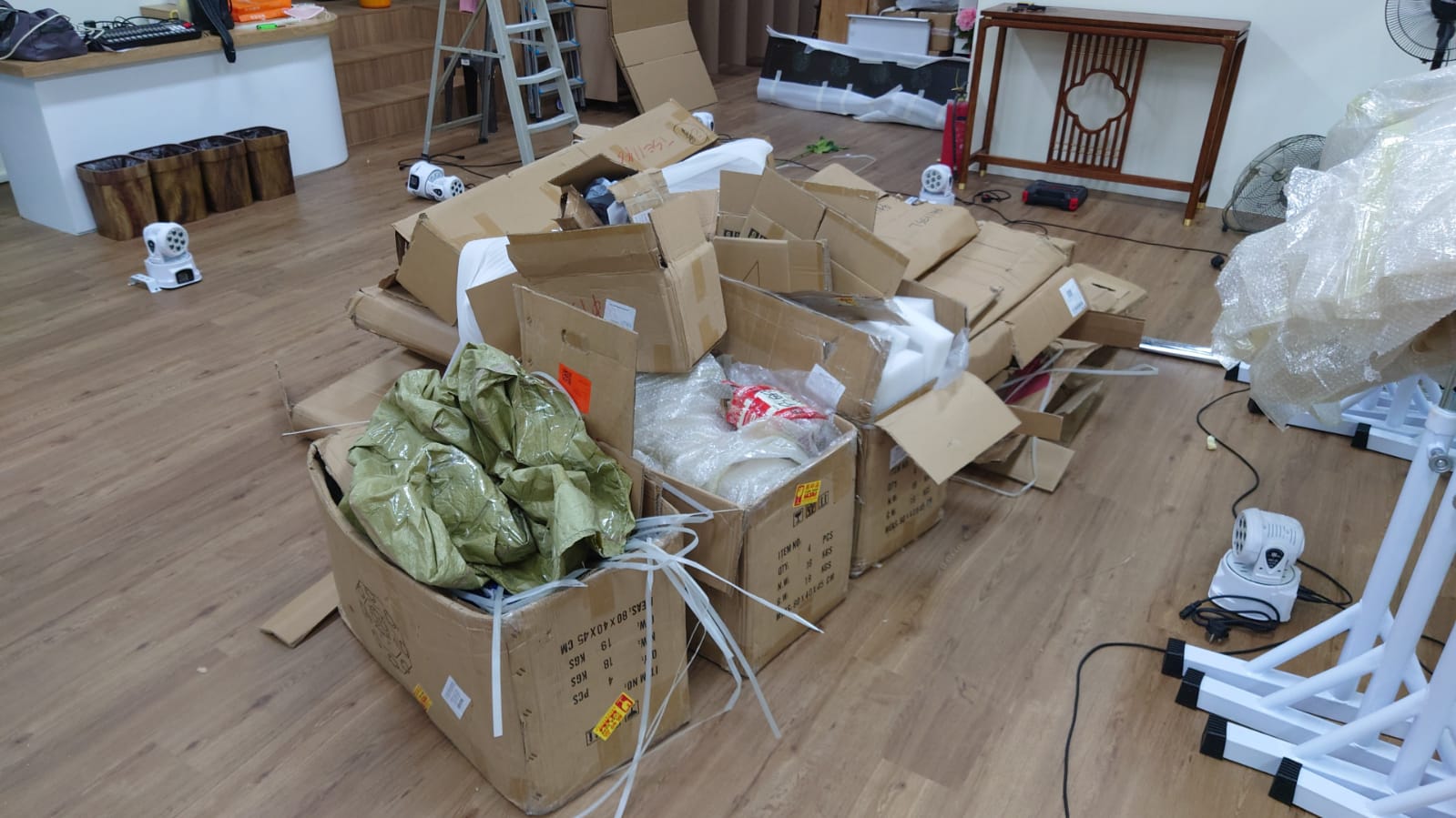 Cardboard boxes and household items stacked on a wooden floor, with some belongings wrapped in green bags.