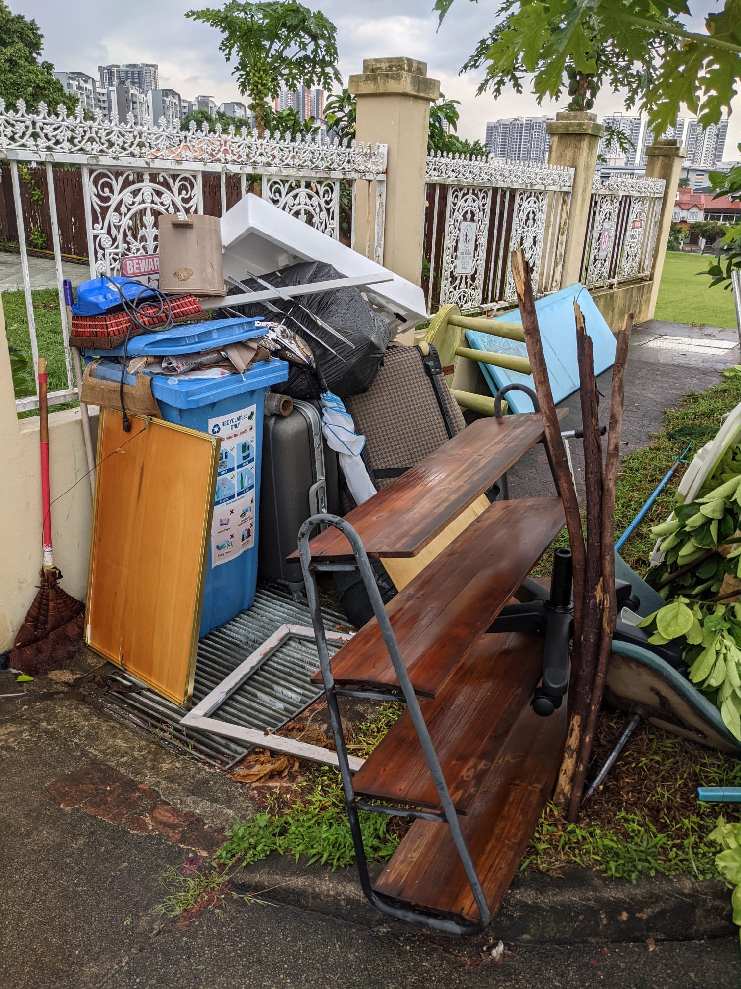 Pile of discarded household items stacked against a fence, including a wooden shelf, blue plastic bin, and other furniture waiting for disposal.