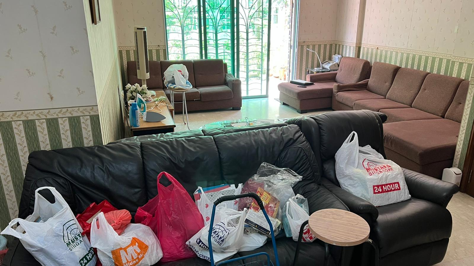 Living room with brown sectional and black leather sofa surrounded by stacked shopping bags and clutter.