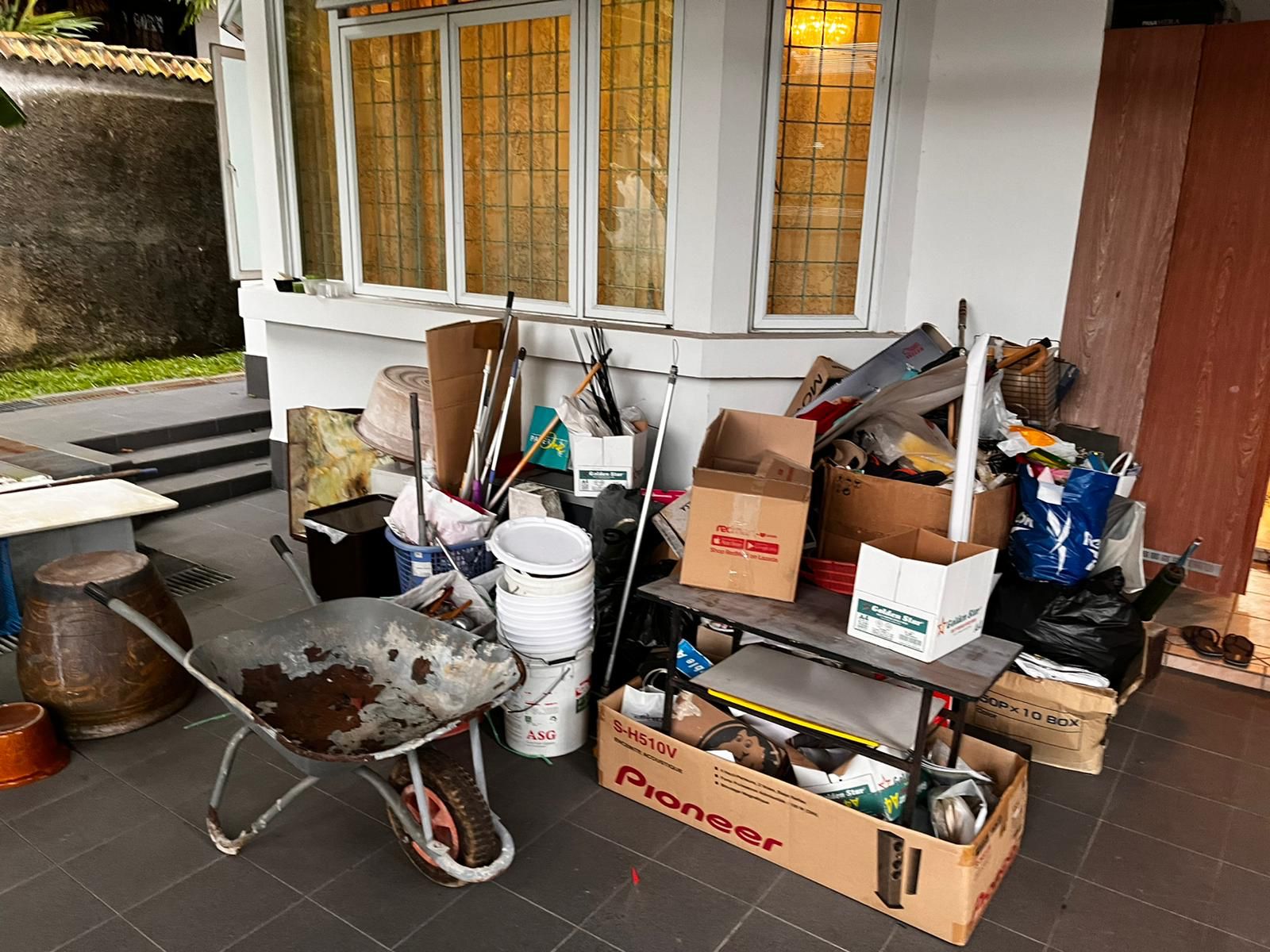 Residential junk pile outside a house including a wheelbarrow, plastic buckets, and flattened cardboard boxes.