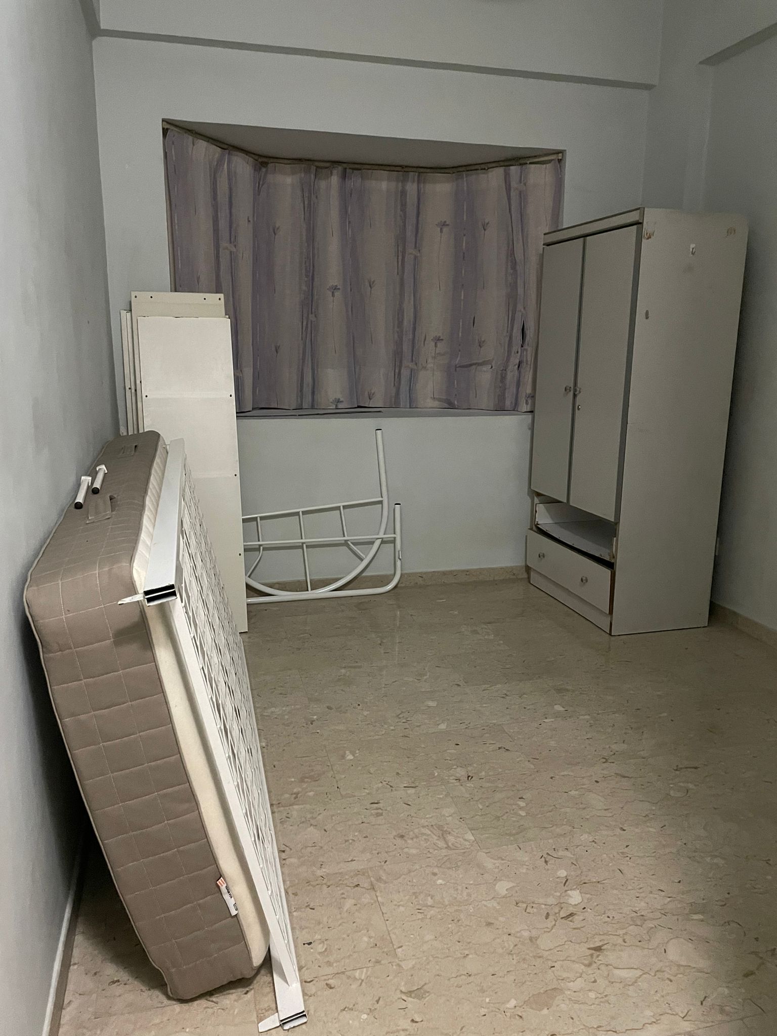 Old mattress propped against a bedroom wall beside a wooden wardrobe and bay window.